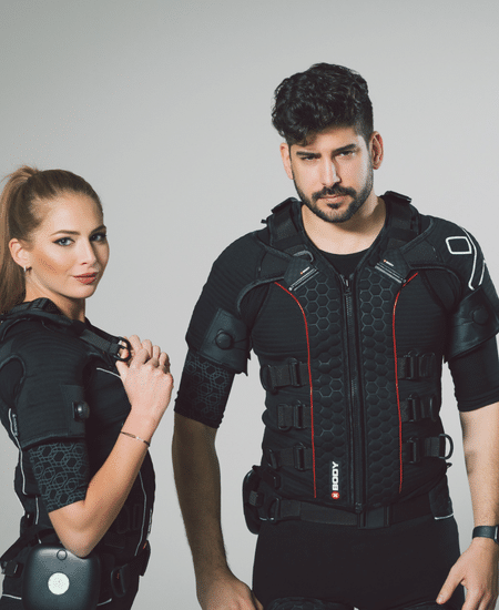 Two people wearing black high-tech suits with hexagonal patterns and shoulder pads stand side by side. The woman adjusts her suit’s strap while the man looks forward. The background is plain and light-colored.