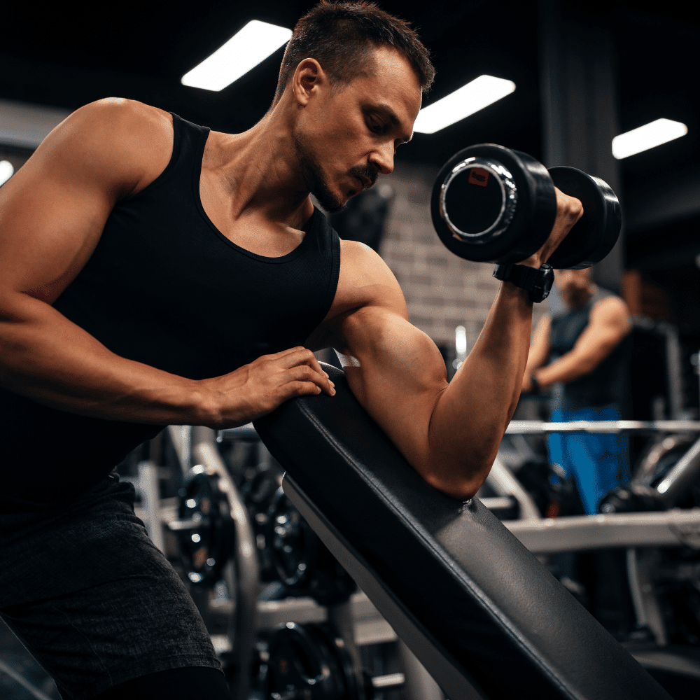 A man in a black tank top performs a bicep curl with a dumbbell in a gym, resting his arm on an incline bench. Fitness equipment and another person are visible in the background.