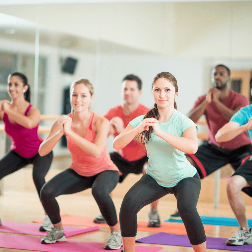 A group of people in athletic wear are doing squats together on yoga mats in a bright fitness studio with mirrored walls.