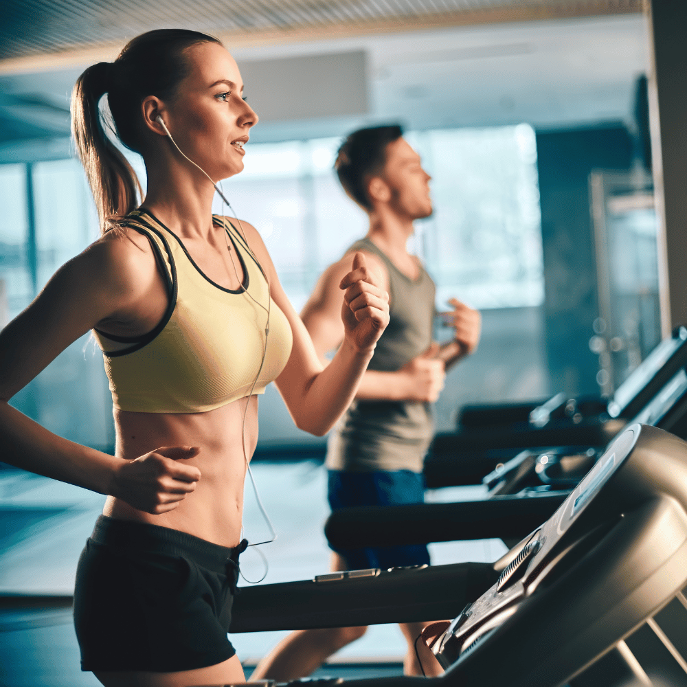 A woman and a man are jogging on treadmills in a gym. The woman is wearing a yellow sports bra and black shorts, listening to earphones, while the man is in the background also running. The setting is bright and modern.