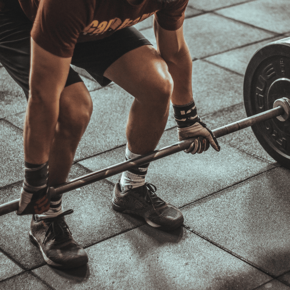 A person wearing gloves, shorts, and sneakers prepares to lift a barbell loaded with weights in a gym, kneeling on a rubber gym floor.