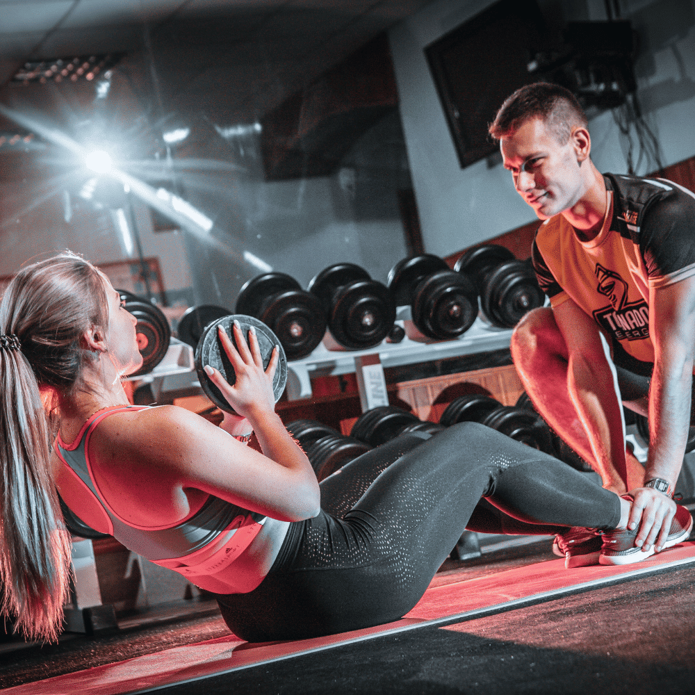 A woman in workout clothes does Russian twists holding a medicine ball, while a male trainer kneels beside her, supporting her feet in a gym with dumbbells in the background.