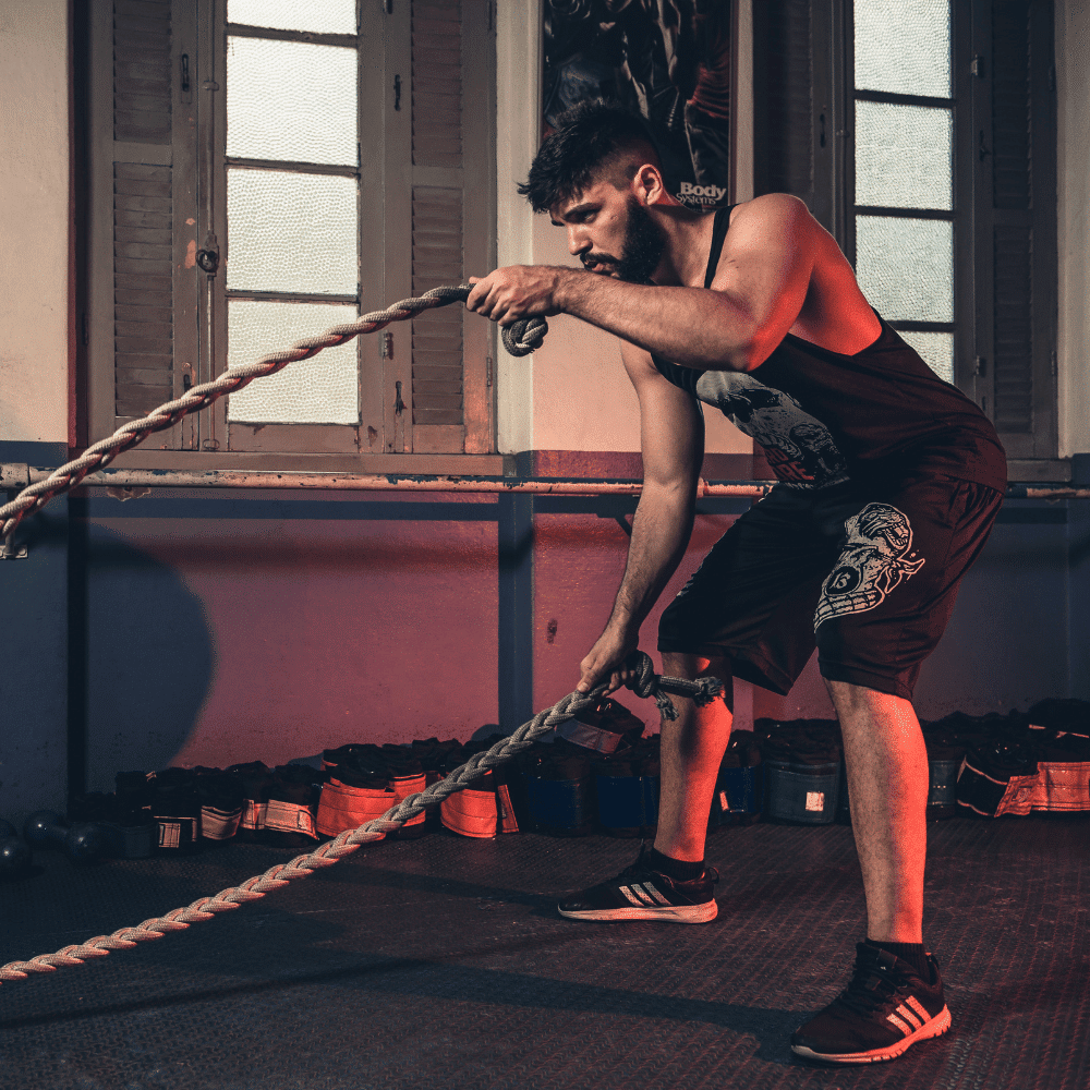 A man in athletic wear exercises indoors with battle ropes, holding each rope in one hand and making a wave motion. He is in a bent-over stance, focused, with gym equipment in the background.