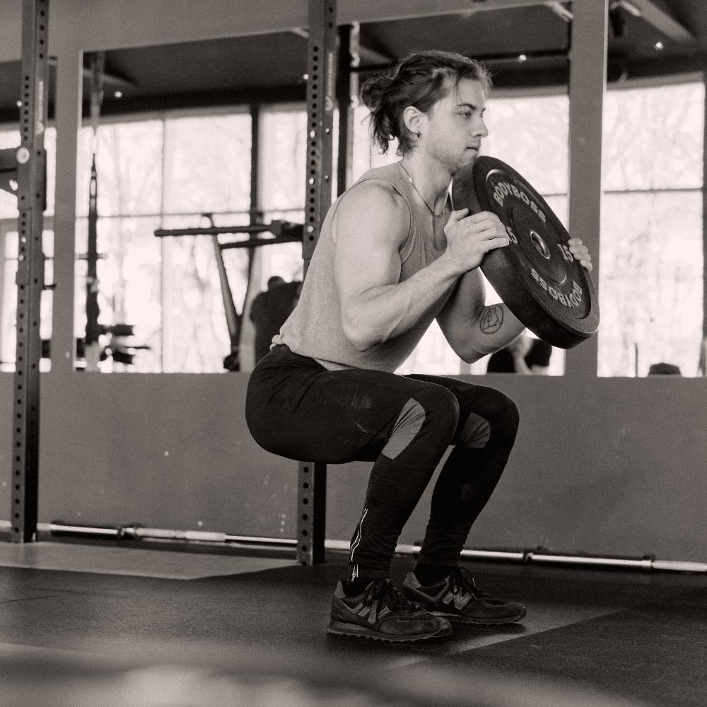 A person in athletic clothing performs a squat while holding a weight plate at chest level in a gym setting with exercise equipment in the background.