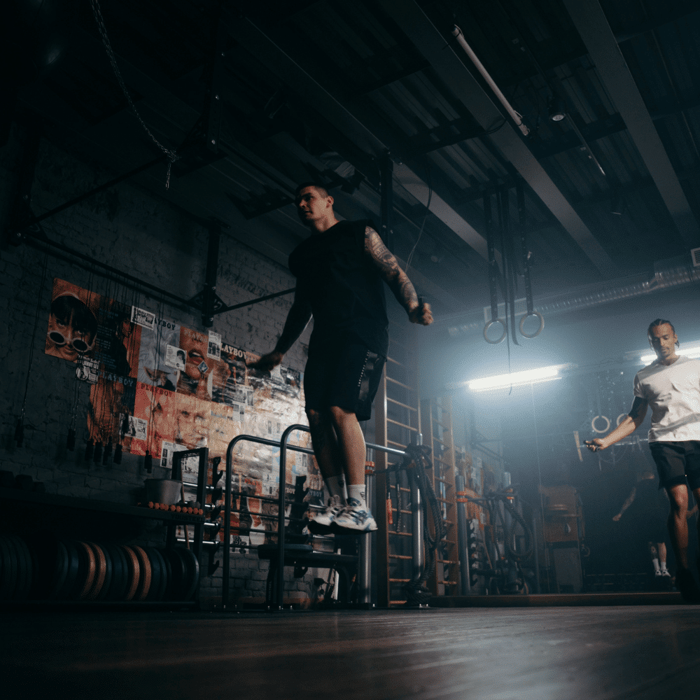 Two men are jump-roping in a dimly lit gym, surrounded by fitness equipment and posters on the brick wall, creating an intense workout atmosphere.