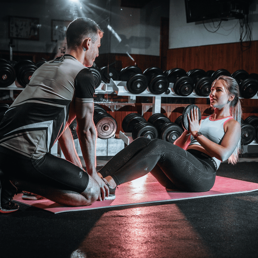 A woman does Russian twists with a weight on a mat in a gym, while a man kneels and holds her feet. Dumbbells are lined up on a rack behind them. Both wear athletic clothing.