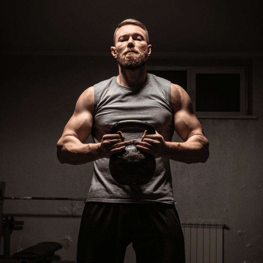 Muscular man with a beard wearing a sleeveless shirt stands indoors, holding a kettlebell with both hands at chest level, and looks confidently at the camera. The background is dimly lit.
